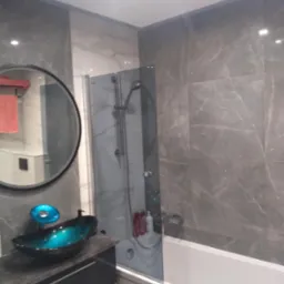 Nowoczesna łazienka z szarymi płytkami imitującymi marmur, owalnym lustrem z czarną ramą, szklaną umywalką w odcieniach turkusu i czerni oraz białą wanną za przeszklonym parawanem.