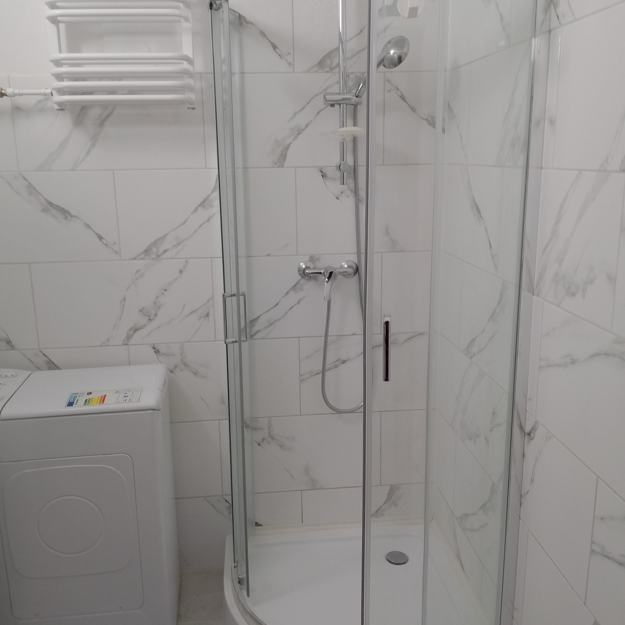 Wykończona łazienka z kabiną prysznicową z chromowaną armaturą, białą pralką i grzejnikiem drabinkowym na ścianie wyłożonej białymi płytkami imitującymi marmur.