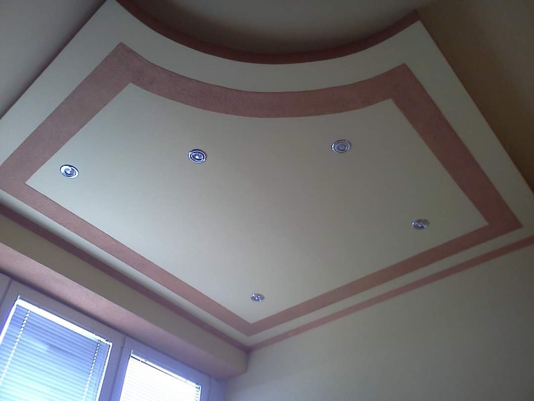Sufit podwieszany z oświetleniem punktowym i dekoracyjnym obramowaniem w kolorze pudrowego różu, widok od dołu, fragment okna z żaluzjami w lewym dolnym rogu.