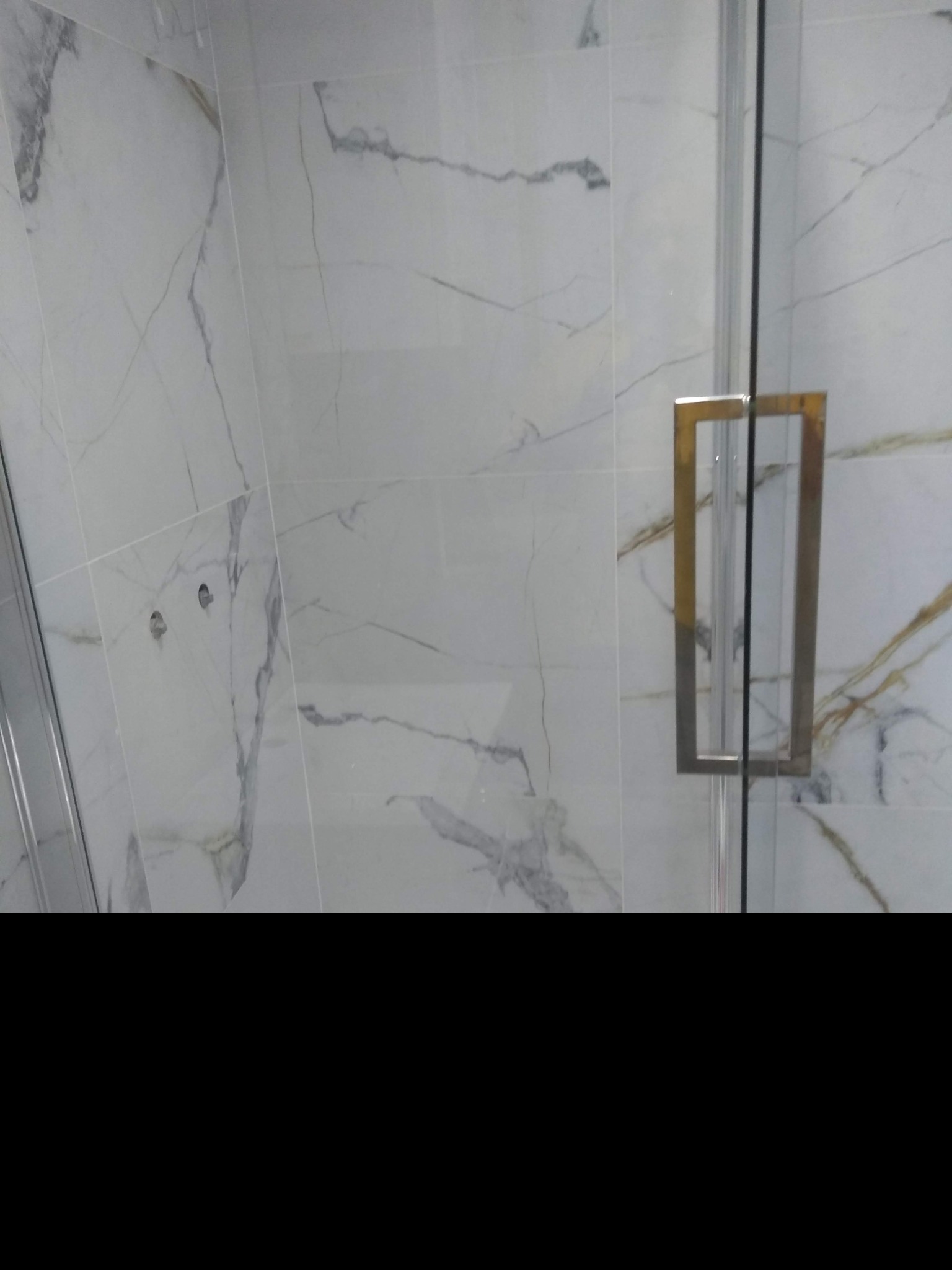 Fragment kabiny prysznicowej z marmurowymi płytkami i złotym uchwytem, widoczne otwory montażowe na ścianie.