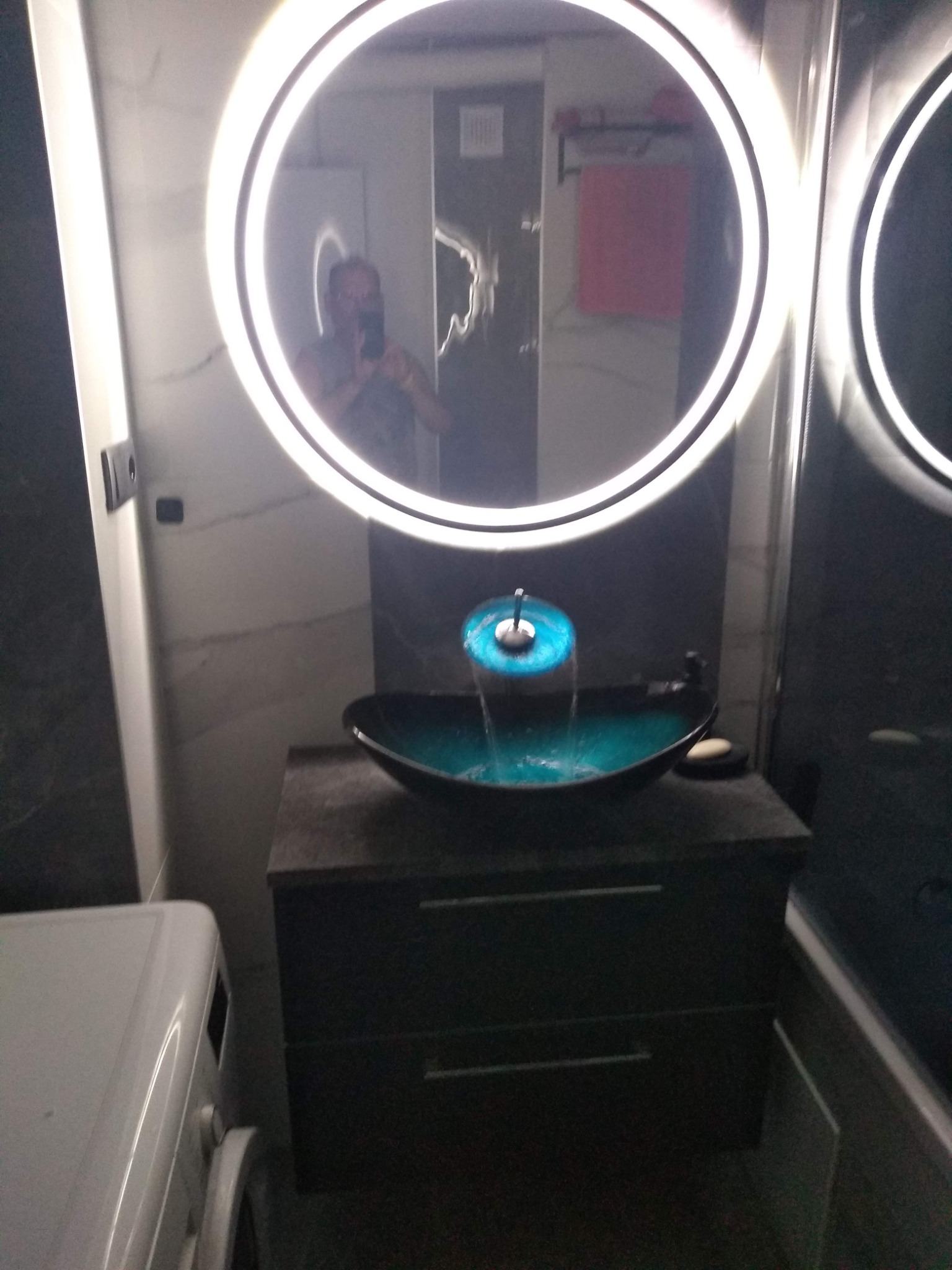 Nowoczesna umywalka z podświetlaną wodą w łazience z okrągłym lustrem LED, widoczny fragment pralki z lewej strony.