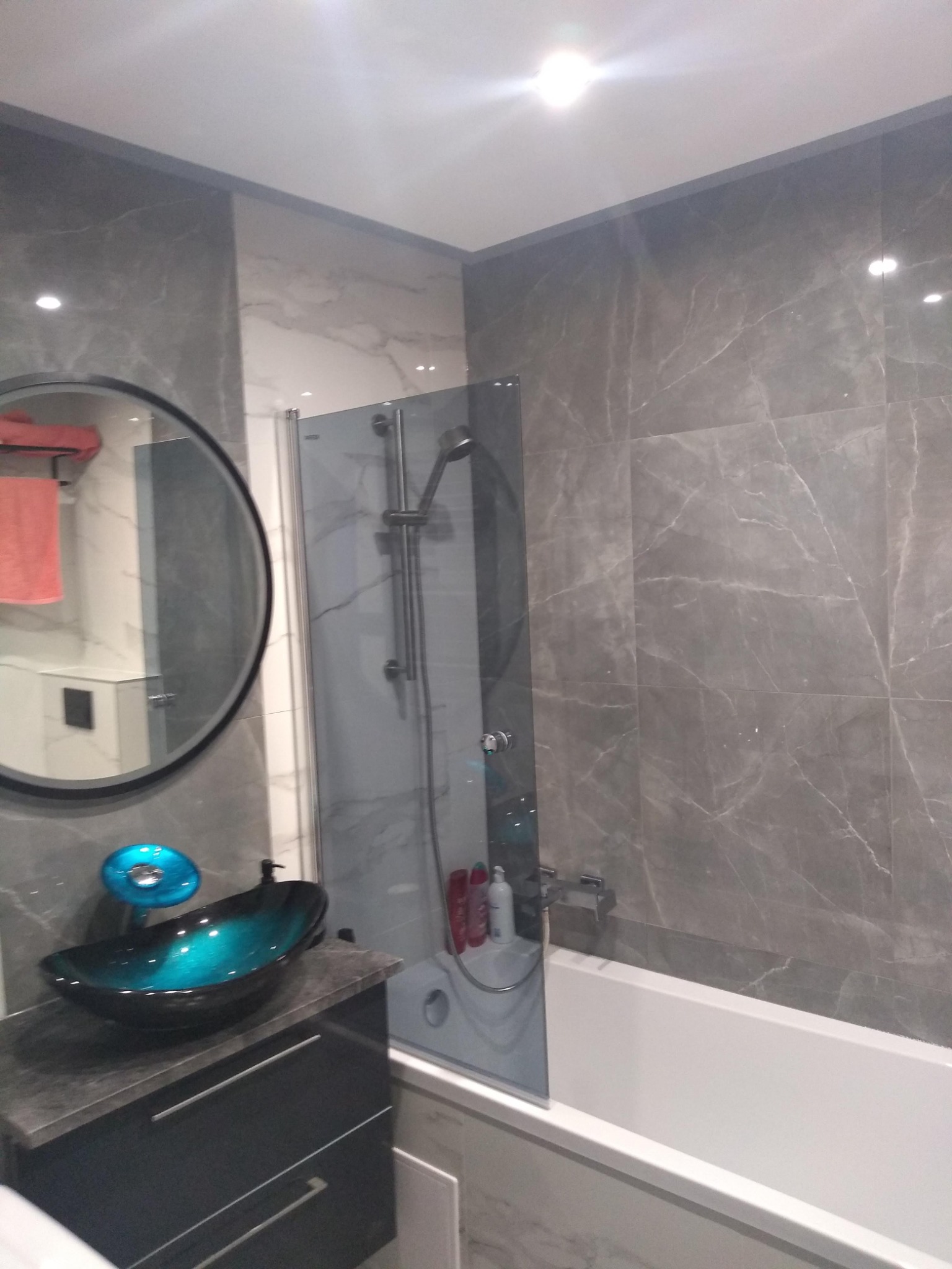 Nowoczesna łazienka z szarymi płytkami imitującymi marmur, owalnym lustrem z czarną ramą, szklaną umywalką w odcieniach turkusu i czerni oraz białą wanną za przeszklonym parawanem.