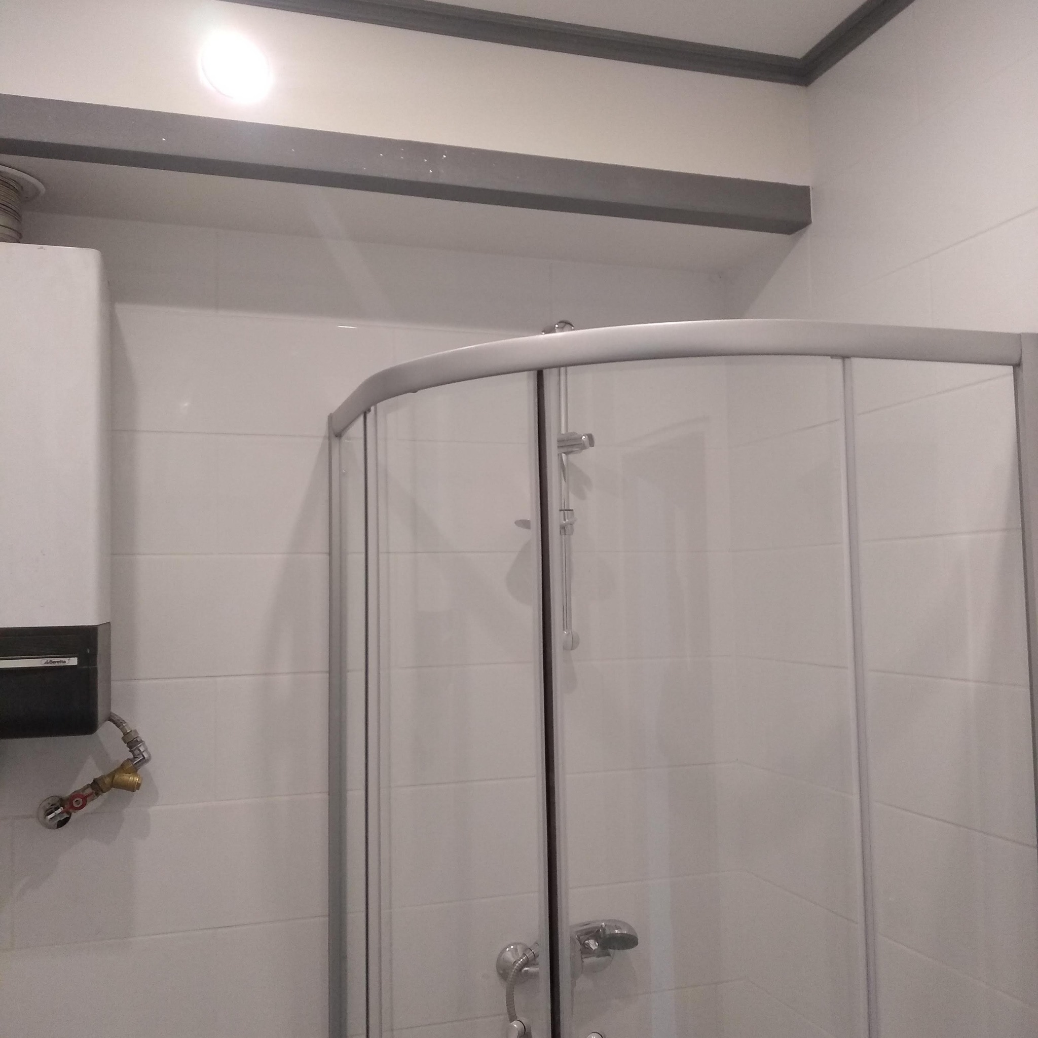 Fragment łazienki z kabiną prysznicową o zaokrąglonych kształtach, białe płytki na ścianach i szara listwa podsufitowa, widoczny bojler z instalacją hydrauliczną