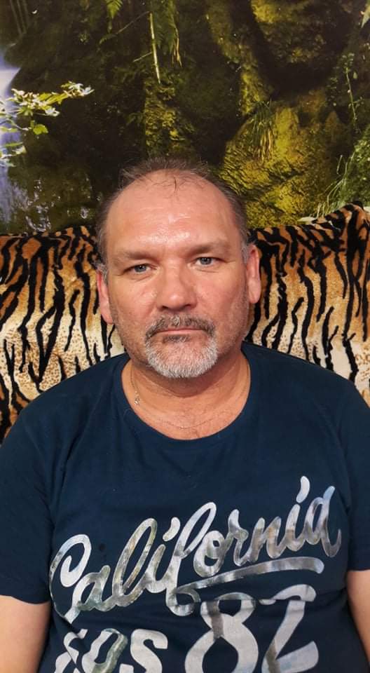 Portret mężczyzny z brodą w niebieskiej koszulce z napisem 'California' na tle wzorzystej tapety imitującej tygrysią skórę i zielonego lasu.