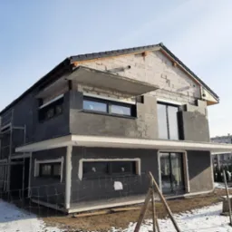 Dwukondygnacyjny dom w trakcie budowy, z widoczną surową konstrukcją z betonu komórkowego i ociepleniem, z częściowo zamontowanymi oknami, na tle zimowego krajobrazu i innych budynków.