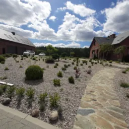 Widok na dwa budynki mieszkalne z czerwonej cegły z szarymi dachami, połączone ścieżką z kamieni naturalnych, otoczone ogrodem z kamieniami i niską roślinnością ozdobną, na tle błękitnego nieba...