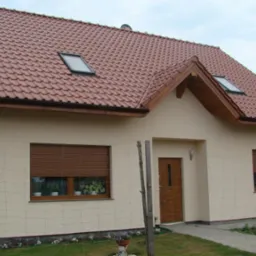 Jasny, otynkowany dom jednorodzinny z czerwoną dachówką, oknami dachowymi i brązowymi roletami w oknach. Widoczny fragment ogrodzenia i trawnik.