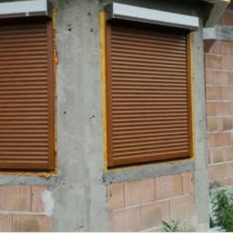 Daniel Jasic - Brązowe rolety zewnętrzne zamontowane w oknach budynku w trakcie budowy, widoczne surowe ściany z betonu komórkowego i pianka montażowa wokół ram okiennych.