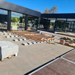 Przygotowanie do ułożenia kostki brukowej na tarasie budynku z dużymi oknami, widoczne drewniane legary, paleta z kostką, narzędzia i piła.