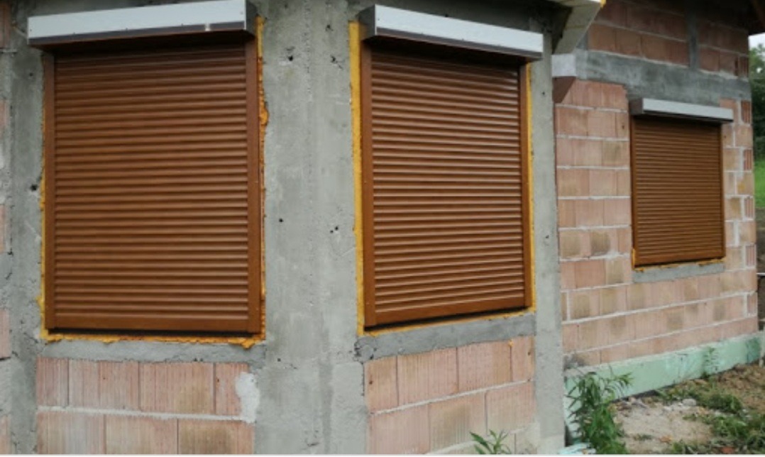 Brązowe rolety zewnętrzne zamontowane w oknach budynku w trakcie budowy, widoczne surowe ściany z betonu komórkowego i pianka montażowa wokół ram okiennych.