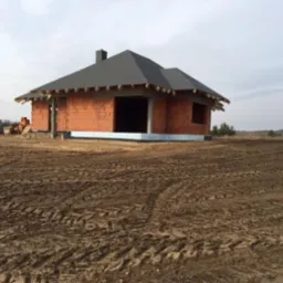 Surowy dom z czerwonej cegły pod szarym dachem na nierównym terenie. Widoczne ślady opon. Częściowo ukończona konstrukcja budynku.