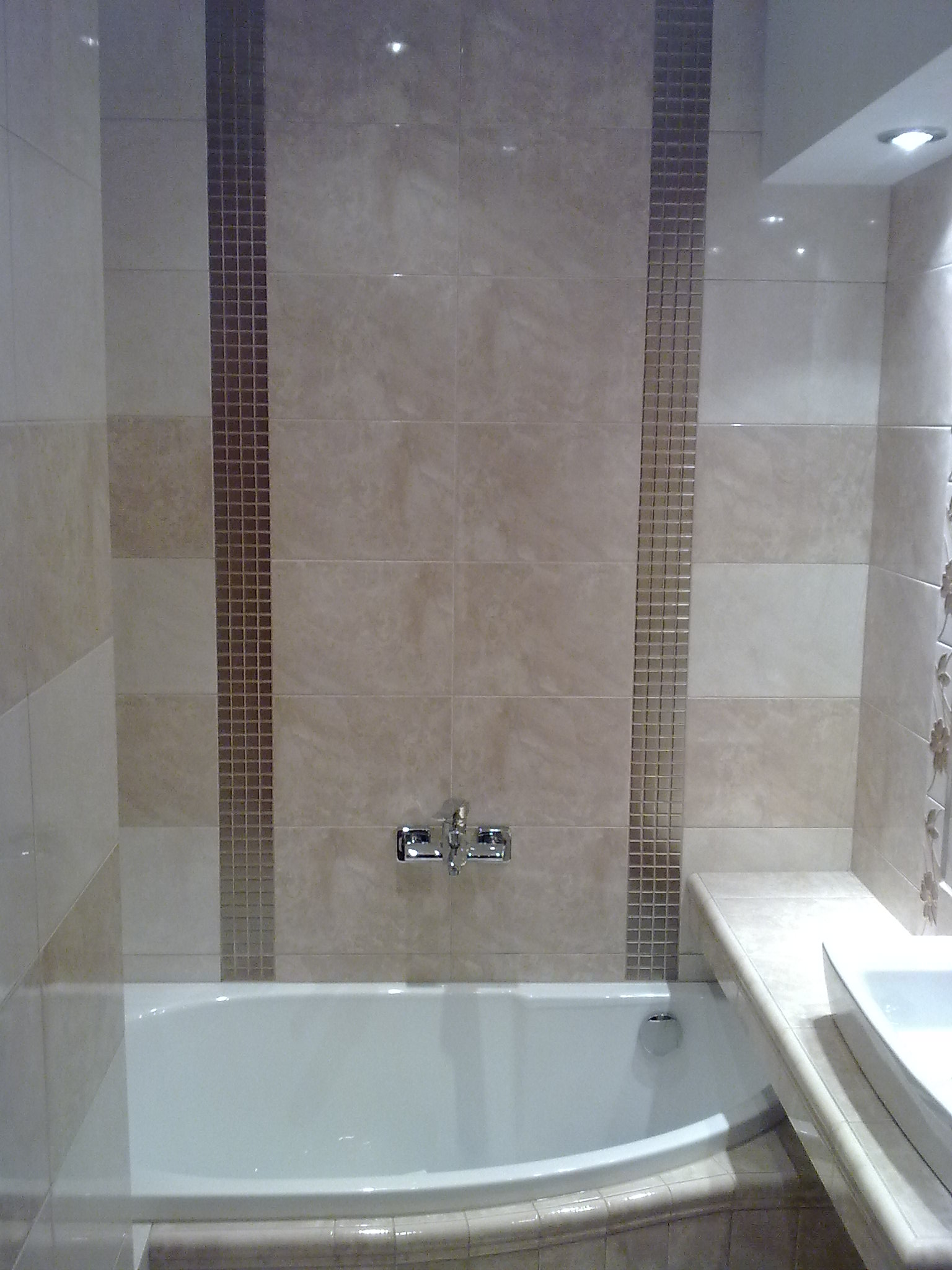 Wykończona łazienka w Gniewkowie z beżowymi płytkami, dekoracyjnymi pasami mozaiki i białą wanną.