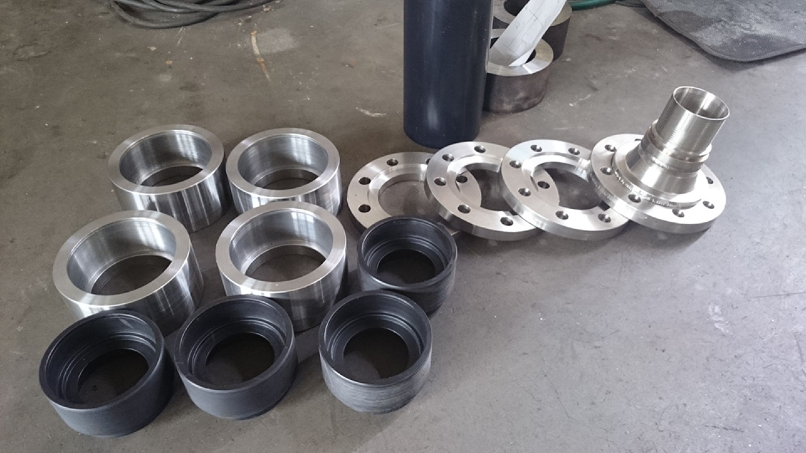 Metalowe elementy: pierścienie (srebrne i czarne), kołnierze z otworami i gwintowany element. Szary beton w tle, widoczne kable. Obróbka metalu.