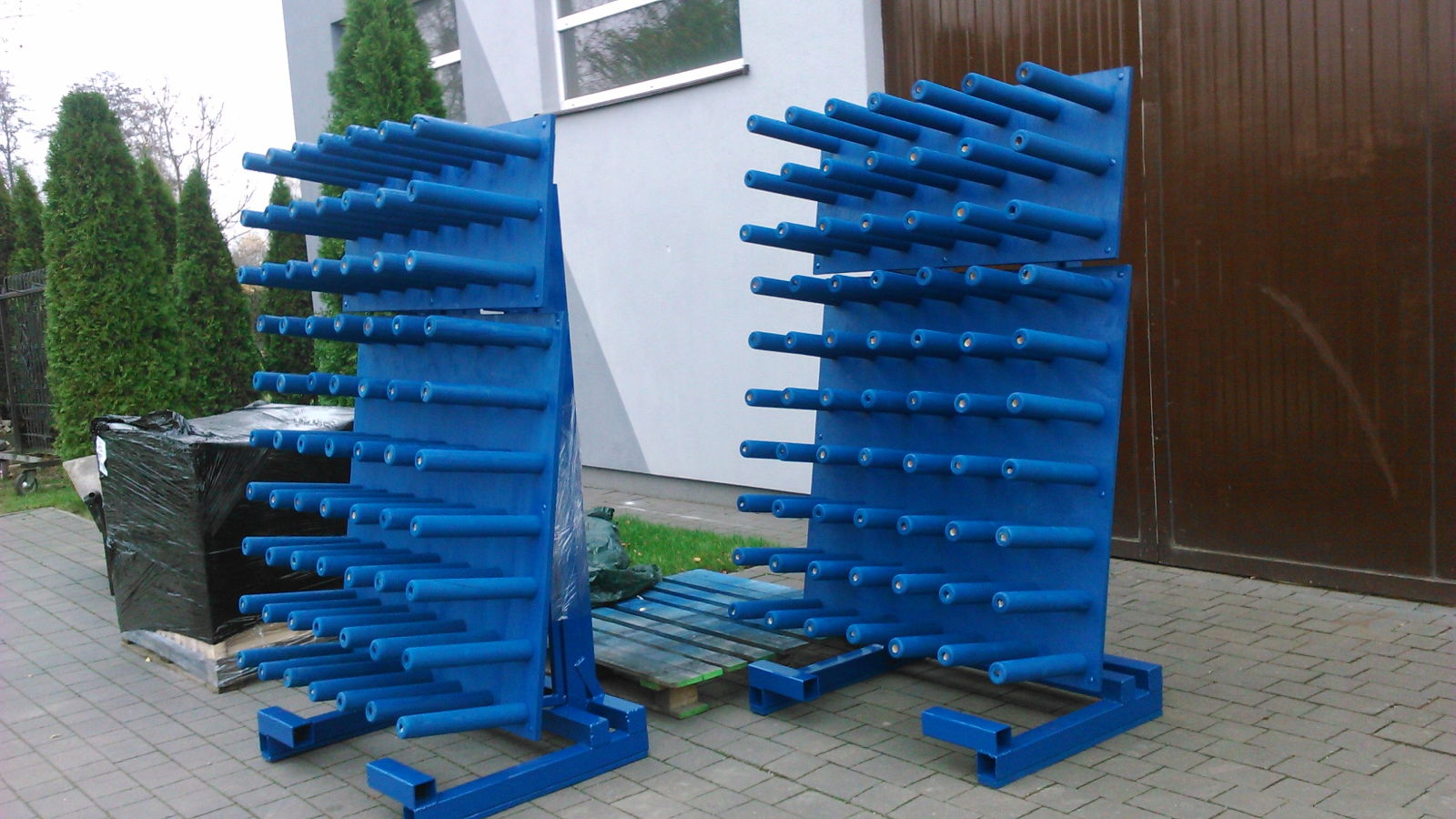 Dwa niebieskie stojaki na elementy metalowe, z licznymi, poziomymi bolcami, na zewnątrz budynku. Widoczne elementy konstrukcyjne i spawane połączenia.