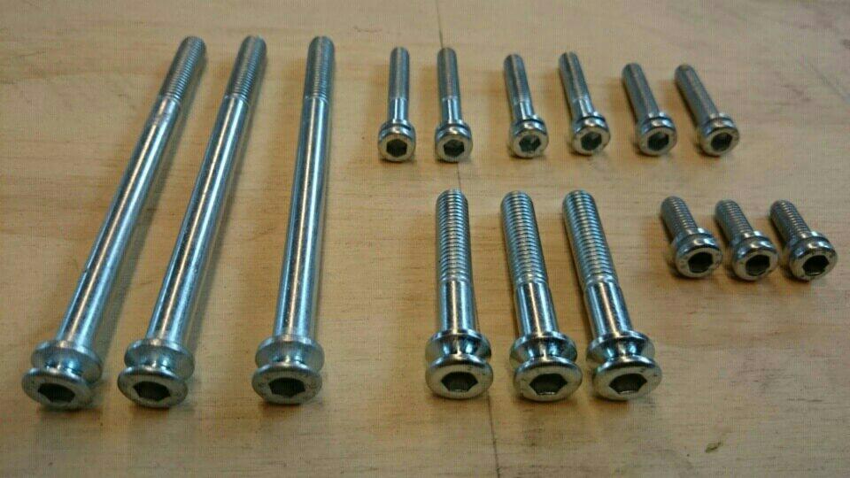 Zestaw srebrnych śrub imbusowych o różnych długościach, ułożonych rzędami na drewnianej powierzchni. Widoczne gwinty i sześciokątne gniazda.