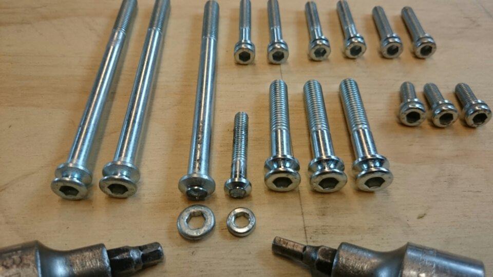 Różnej wielkości śruby i podkładki ze stali, ułożone na drewnianym blacie, z widocznymi narzędziami w dolnej części kadru. Precyzyjne wykonanie detali.