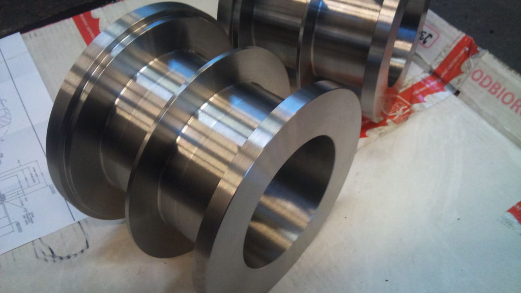 Precyzyjnie wykonane, metalowe pierścienie z rowkami, leżące na dokumentacji technicznej i kartonie, prezentują detale obróbki. Metaliczny połysk, dokładność wykonania.