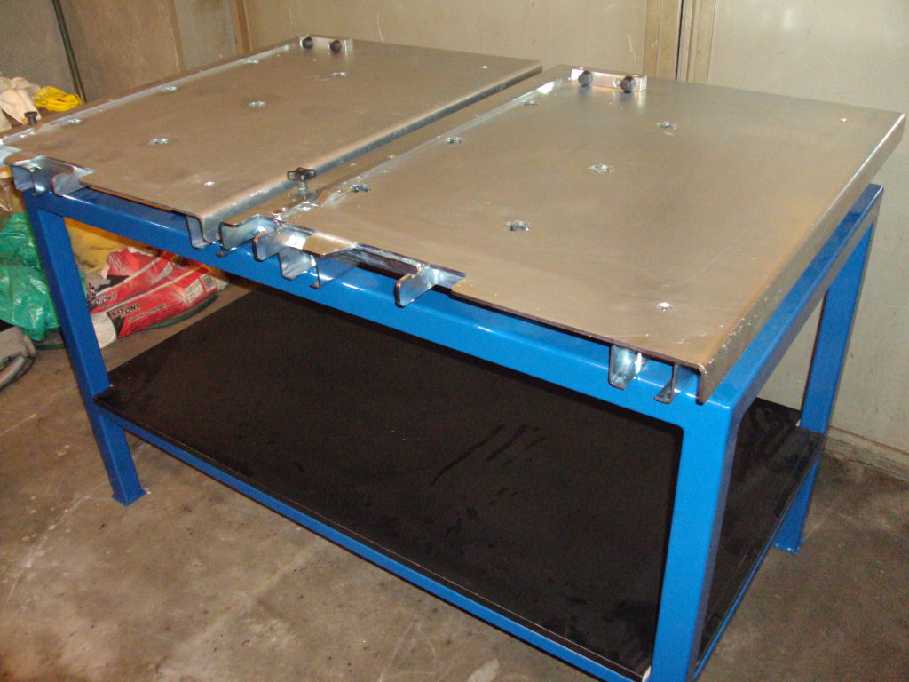 Niebieski stół warsztatowy z metalowym blatem i dolną półką. Blat składa się z dwóch części, z widocznymi otworami i elementami mocującymi.