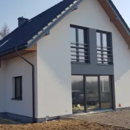 Nowoczesny dom jednorodzinny z białą elewacją, ciemną dachówką i antracytowymi oknami, widok z zewnątrz na świeżo zagospodarowany teren wokół budynku.