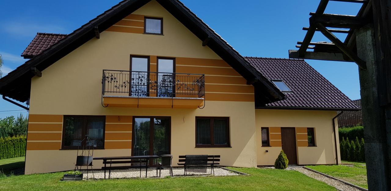 Dwukondygnacyjny dom z beżową elewacją i ciemnobrązowymi akcentami, dekoracyjny balkon z kutą balustradą, zadbany trawnik z elementami małej architektury ogrodowej.