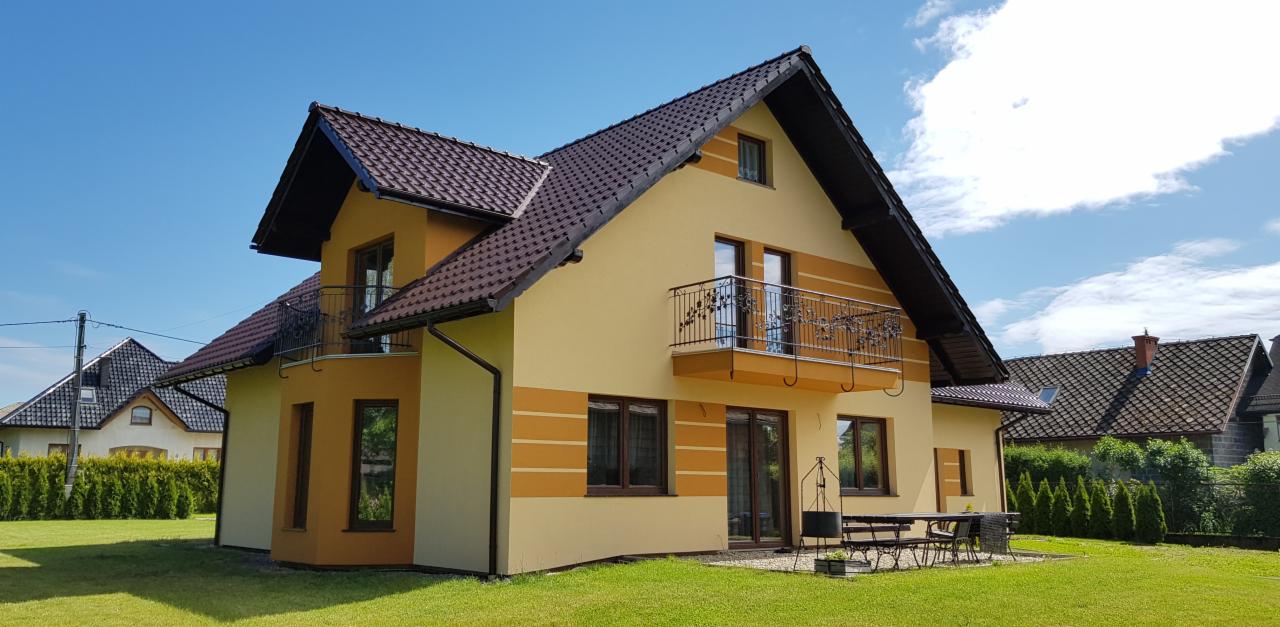 Dwukondygnacyjny dom z jasną elewacją w odcieniach beżu i brązu, z balkonami z ozdobnymi balustradami, brązową dachówką i widocznym fragmentem ogrodu z meblami ogrodowymi.