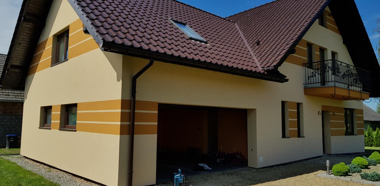 Dom jednorodzinny z beżową elewacją ozdobioną poziomymi pasami w kolorze ochry, brązowy dach z dachówki ceramicznej, balkon z kutą balustradą, widok z zewnątrz w słoneczny dzień.