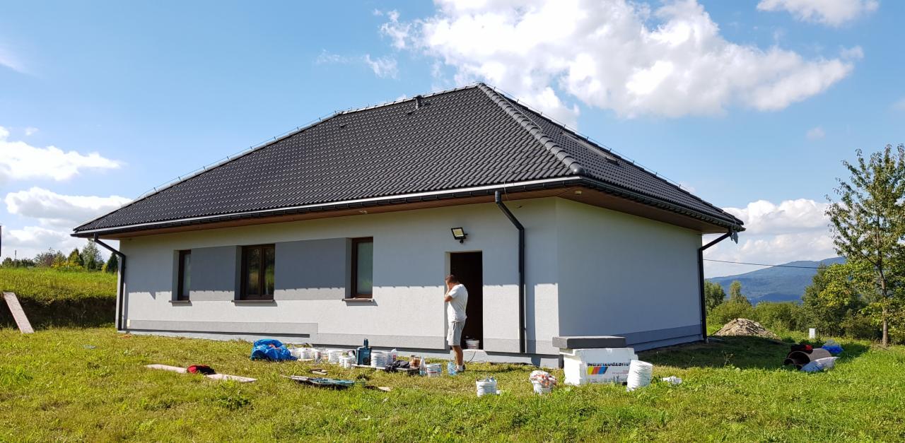 Jednopiętrowy dom z szarą elewacją w trakcie prac wykończeniowych, widoczne puszki z farbą i pracownik przy wejściu, w tle górzysty krajobraz.