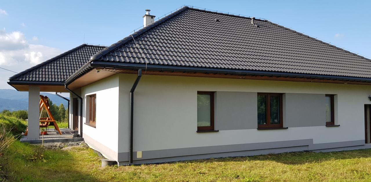 Nowoczesny dom jednorodzinny w Porąbce z jasną elewacją, szarymi akcentami i ciemną dachówką, widok z boku na tle górskiego krajobrazu.