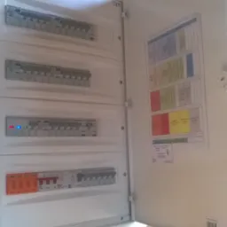 Otwarta skrzynka elektryczna z bezpiecznikami na ścianie z czerwonej cegły, widoczne kable w peszlach i schemat instalacji elektrycznej wewnątrz drzwiczek.