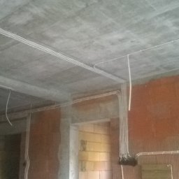 Instalacje Elektryczne Bogdan Zęgota - Wnętrze surowego domu w trakcie prac instalacyjnych: widoczne kable elektryczne prowadzone po suficie i ścianach, ceglane ściany w stanie deweloperskim, brak wykończenia.