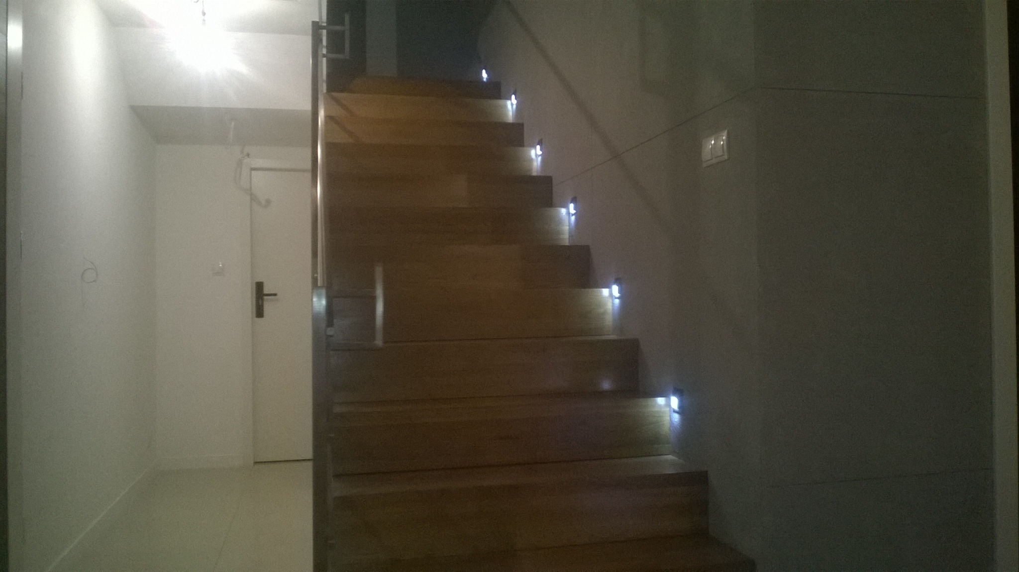 Drewniane schody z zamontowanym oświetleniem LED na każdym stopniu, w tle biała ściana i drzwi.