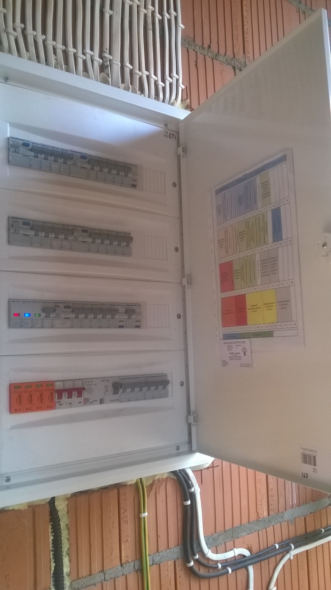 Otwarta skrzynka elektryczna z bezpiecznikami na ścianie z czerwonej cegły, widoczne kable w peszlach i schemat instalacji elektrycznej wewnątrz drzwiczek.