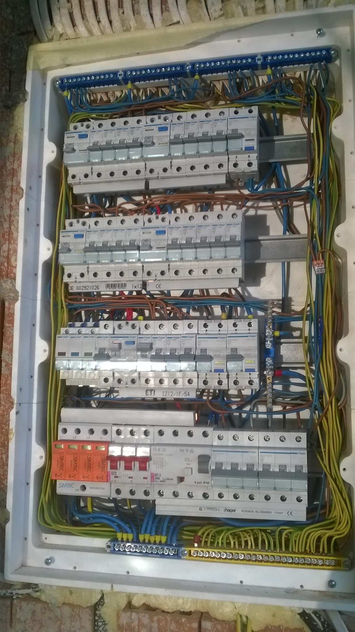 Rozdzielnica elektryczna z zabezpieczeniami przepięciowymi Simtec, widoczne liczne przewody w różnych kolorach, zamontowana w białej obudowie na tle muru z cegieł.