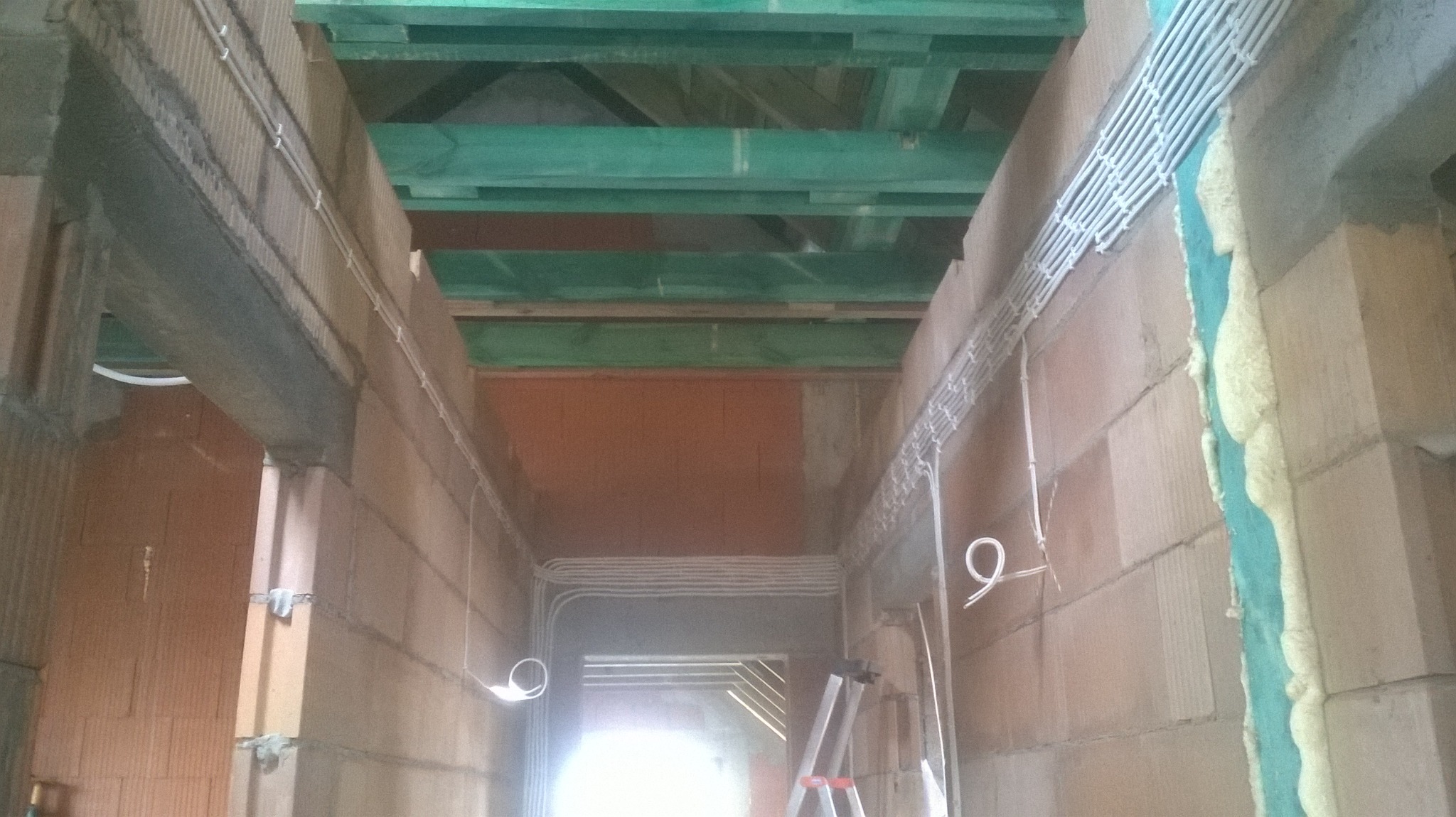 Wnętrze w trakcie budowy z widocznymi przewodami elektrycznymi ułożonymi w korytach na ścianach z bloczków, zielone belki stropowe, pianka montażowa w szczelinie i drabina aluminiowa.