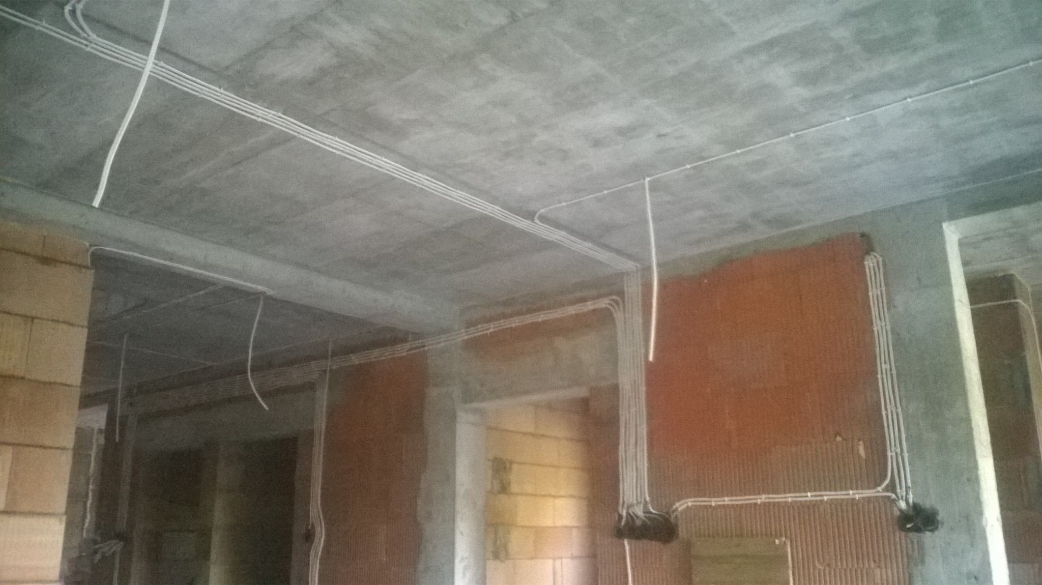 Wnętrze surowego domu w trakcie prac instalacyjnych: widoczne kable elektryczne prowadzone po suficie i ścianach, ceglane ściany w stanie deweloperskim, brak wykończenia.