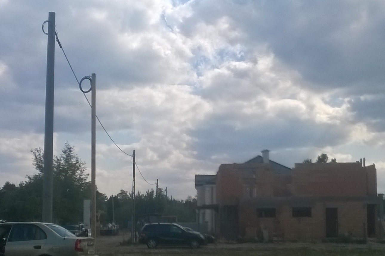 Słupy energetyczne z przewodami zasilającymi nowy, murowany dom w trakcie budowy, widoczne samochody i pochmurne niebo.