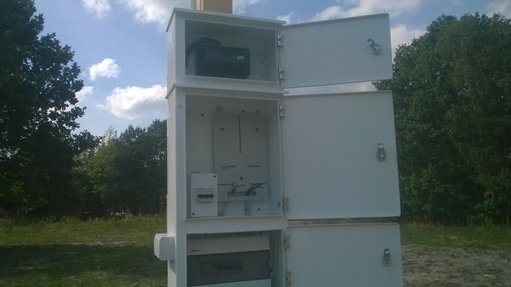 Biała szafka elektryczna z otwartymi drzwiczkami na tle drzew i nieba, widoczne zabezpieczenia, przewody i zasilacz awaryjny