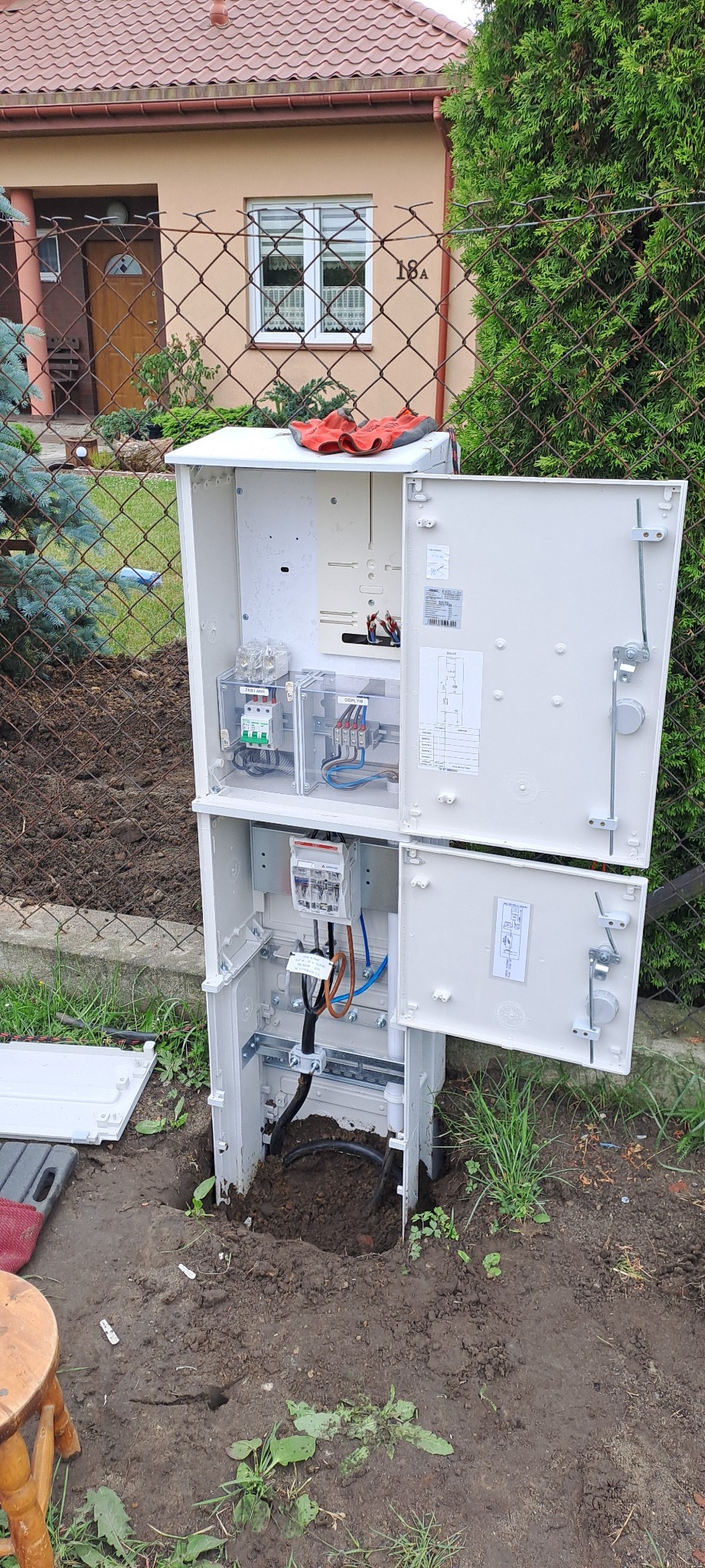 Otwarta, dwuczęściowa skrzynka elektryczna zamontowana w ziemi przed domem, widoczne wewnętrzne okablowanie i zabezpieczenia, na górnej części leżą robocze rękawice.