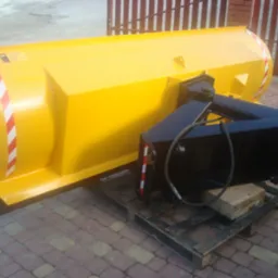 Żółty, nowy pług do śniegu z czarnym mocowaniem, umieszczony na drewnianej palecie, z widocznymi pasami ostrzegawczymi.