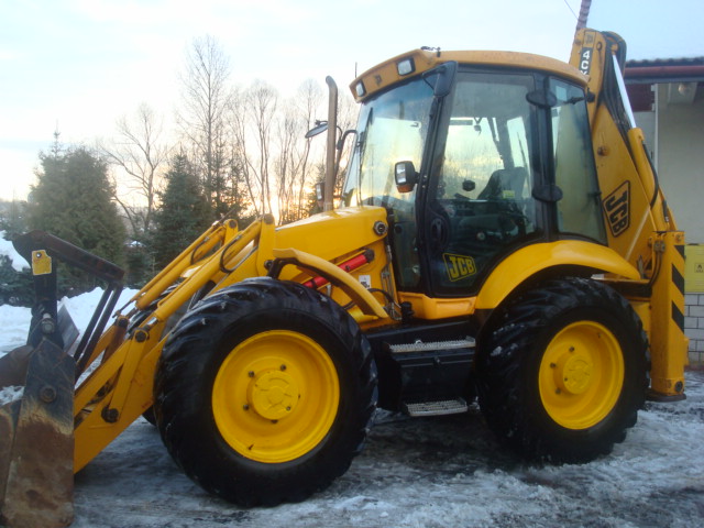 Żółta koparko-ładowarka JCB 4CX zaparkowana na śniegu przed budynkiem w zimowy dzień. Widoczne logo JCB na kabinie i ramieniu koparki.