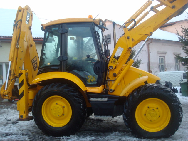 Żółta maszyna budowlana JCB z widocznym ramieniem koparki i ładowarki, zaparkowana na placu z resztkami śniegu, z budynkami w tle.