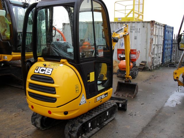 Żółta mini koparka JCB 8018 z kabiną, widoczna z przodu, na placu budowy z kontenerami w tle.