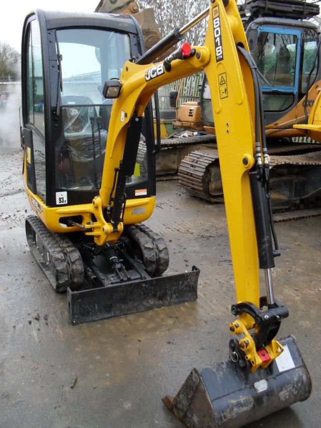 Żółta mini koparka JCB 8018 z czarną kabiną i łyżką, stojąca na mokrym placu, z inną maszyną budowlaną w tle.