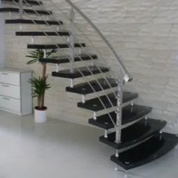 Nowoczesne schody z czarnymi stopniami i nierdzewną balustradą, wsparte na metalowych wspornikach, na tle ściany z dekoracyjnej białej cegły, obok biała komoda i roślina doniczkowa.