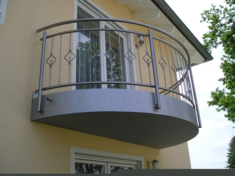 Półokrągły balkon z balustradą ze stali nierdzewnej, zdobioną romboidalnymi elementami, zamontowany na elewacji budynku w jasnym odcieniu beżu.