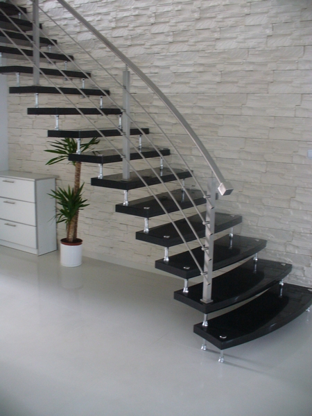 Nowoczesne schody z czarnymi stopniami i nierdzewną balustradą, wsparte na metalowych wspornikach, na tle ściany z dekoracyjnej białej cegły, obok biała komoda i roślina doniczkowa.