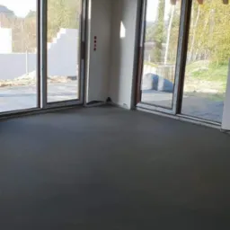 Surowe wnętrze domu w trakcie budowy z dużymi oknami przesuwnymi i wylaną posadzką.