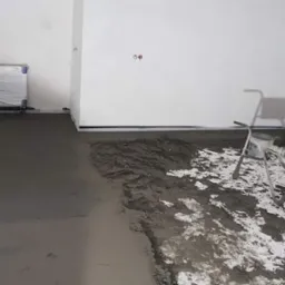 Wnętrze w trakcie wylewania posadzki: świeży, szary beton częściowo rozprowadzony, widoczny agregat do wylewek i zabezpieczony folią grzejnik.