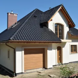 Nowy dom jednorodzinny z ciemną, błyszczącą dachówką i jasną elewacją. Widoczny garaż z drewnianą bramą i ceglany komin. Ujęcie w słoneczny dzień.