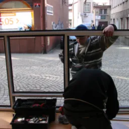 Montaż ramy okiennej z widokiem na brukowaną ulicę; dw&oacute;ch monter&oacute;w w trakcie pracy, jeden przytrzymuje ramę, drugi pracuje nad uszczelnieniem; skrzynki z narzędziami obok.